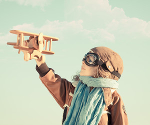 young child dressed as an old-fashioned pilot holing a wooden airplane